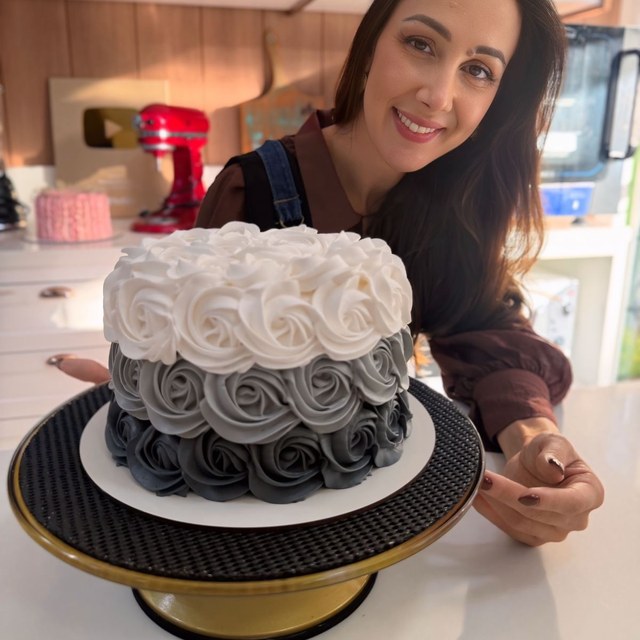 Confeiteira ao lado de bolo decorado com rosetas brancas e cinza